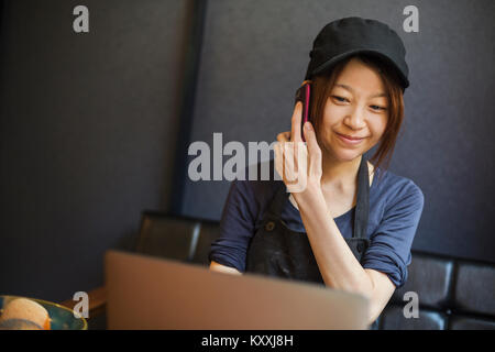 Woman working in a bakery, wearing baseball cap, sitting at table in front of laptop, using mobile phone. Stock Photo