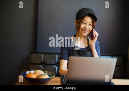 Woman working in a bakery, wearing baseball cap, sitting at table in front of laptop, using mobile phone. Stock Photo