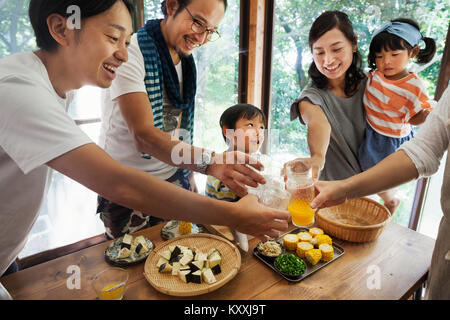 Two men, woman holding young girl and boy gathered around a table with food, holding drinking glasses, toasting. Stock Photo