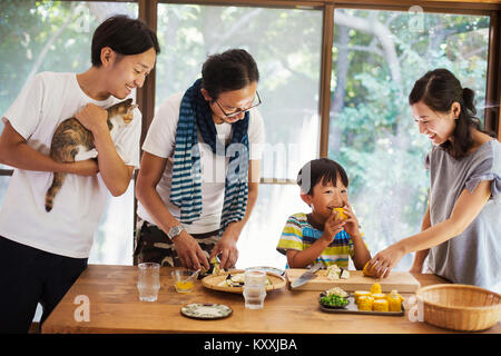 Two men, woman and boy gathered around a table, preparing corn on the cob, one man holding cat, smiling. Stock Photo