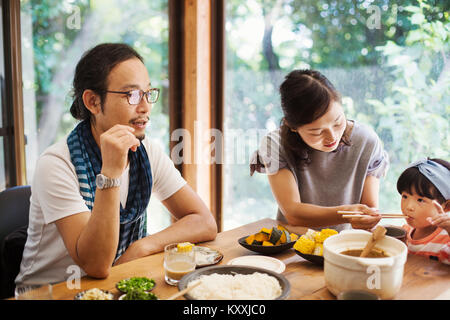 Man, woman and young girl sitting round a table with bowls of food, eating together. Stock Photo