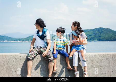Family, man, boy and woman with young girl on her lap sitting side by side on a wall by the ocean. Stock Photo
