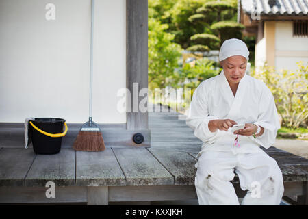 Buddhist monk wearing white robe and cap sitting on wooden floor outside a temple, using mobile phone. Stock Photo