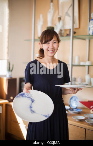 Smiling woman standing in a Japanese porcelain shop, holding two white plates with blue decoration. Stock Photo