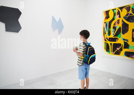 Boy with short black hair wearing backpack standing in art gallery, holding pen and paper, looking at modern painting. Stock Photo