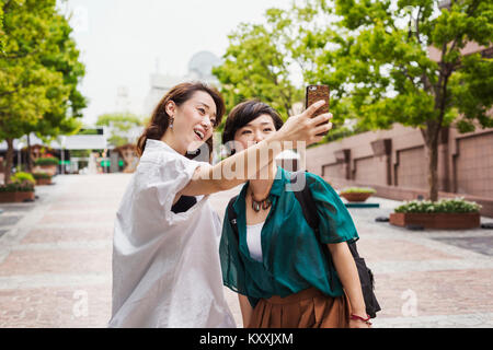 Two women with black hair wearing white and green shirt standing outdoors, taking selfie with mobile phone, smiling. Stock Photo