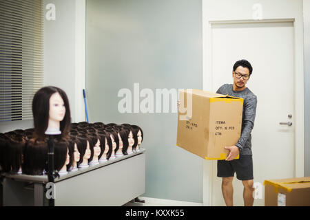 Bearded man wearing glasses standing indoors, carrying cardboard box, large group of mannequin heads with brown wigs on table. Stock Photo
