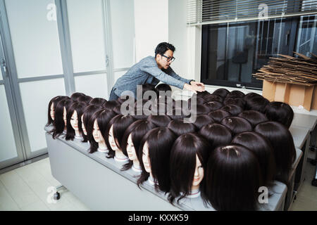 Bearded man wearing glasses standing indoors, arranging mannequin heads with brown wigs. Stock Photo