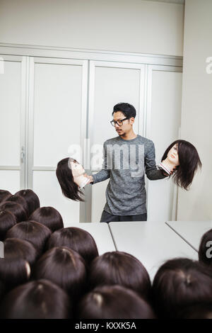 Bearded man wearing glasses standing indoors, holding mannequin head with brown wig in each hand. Stock Photo