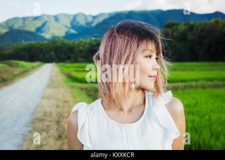 A young woman standing in open space by rice paddy fields of green shoots, and mountain landscape. Stock Photo