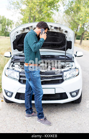 Young man using his cell phone in front of a broken down car parked on the side of a road Stock Photo