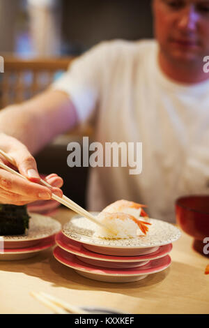 Close up of man sitting at table in Asian restaurant, picking up piece of sushi from a plate with chopsticks. Stock Photo