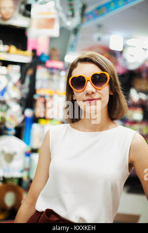Young woman with brown hair wearing white sleeveless top wearing orange heart shaped sunglasses, looking at camera. Stock Photo