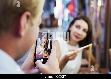 Man taking picture with smartphone of woman sitting at a table in an asian restaurant, using chopsticks, eating noodles. Stock Photo