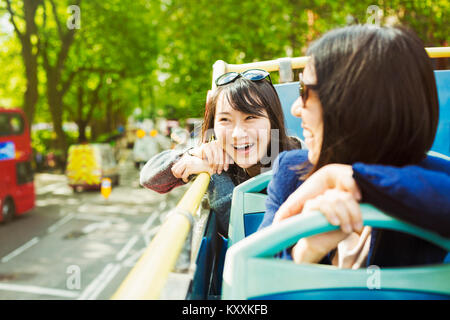 Two smiling women with black hair sitting on the top of an open Double-Decker bus driving along tree-lined urban road. Stock Photo