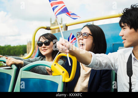 Smiling man waving Union Jack flag and two women with black hair sitting on the top of an open Double-Decker bus. Stock Photo