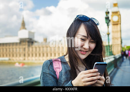 Smiling woman with black hair looking at smartphone, standing on Westminster Bridge over the River Thames, London, with the Houses of Parliament and B Stock Photo