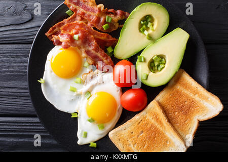 Delicious breakfast of eggs with crispy bacon, avocado, toast and tomatoes close-up on a black plate on the table. Horizontal top view from above