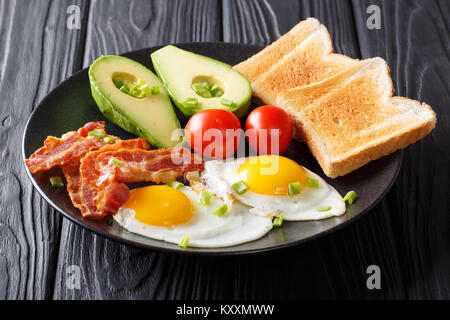 Freshly fried eggs with crispy bacon, avocado, toast and tomatoes close-up on a black plate on a table. Tasty breakfast. horizontal Stock Photo