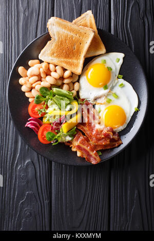 Delicious fried eggs, bacon, beans, toast and fresh vegetable salad on a plate on the table. Vertical top view from above Stock Photo