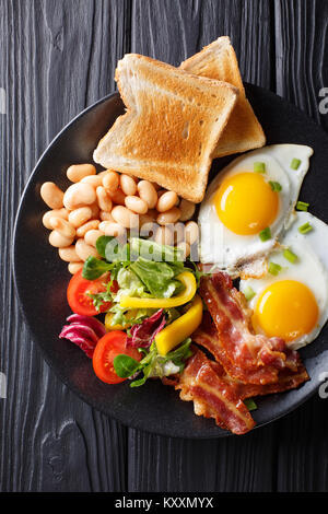 Fresh breakfast: fried eggs with bacon, beans, toast and vegetable salad on a black plate on the table. Vertical top view from above Stock Photo