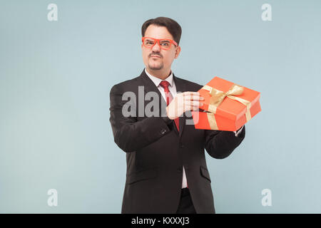 Unhappy man in black suit holding opened red gift box and looking at camera. Studio shot, isolated on light blue background Stock Photo