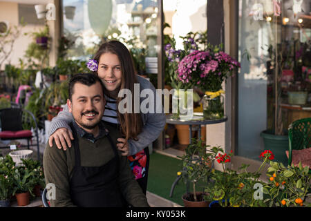 Smiling florist couple in a flower shop. Male wearing black apron sitting in a chair and smiling female with a lisianthus flower in her hair behind hi Stock Photo