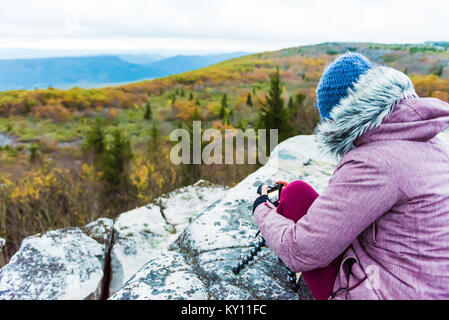Sitting young woman photographer with camera and tripod on cold autumn morning in jacket coat, wool hat in Bear Rocks, West Virginia taking pictures o Stock Photo