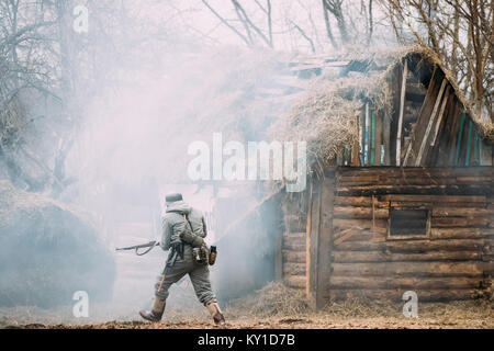 Re-enactor Dressed As German Wehrmacht Infantry Soldier In WW II Running On Battlefield Near Burning Wooden House. Stock Photo