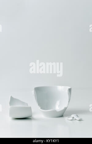 a broken white ceramic coffe cup with its pieces spread out on a white table, against a white background with a blank space on top Stock Photo