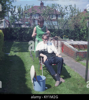 1960s, historical, a woman giving a man sitting in a metal garden seat wearing his slippers a haircut outside on the lawn in a suburban garden, England, UK. Stock Photo