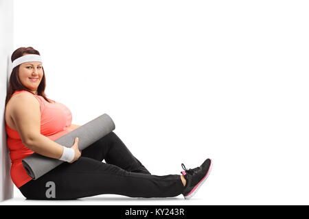 Overweight woman with an exercise mat sitting on the floor and leaning against a wall isolated on white background Stock Photo