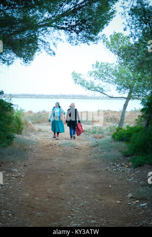 two middle aged women chatting walk away from camera towards an opening in a forest with a lake in the distance Stock Photo