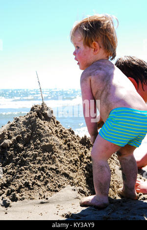 a two year old toddler in swimming trunks stands next to a sand mountain on beach being built by him and his older brother, France Stock Photo