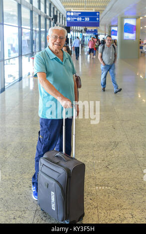 Model Released: Senior man (80-85) smiling while holding a hand luggage at a busy airport terminal - Concept: older passengers happy to be traveling Stock Photo