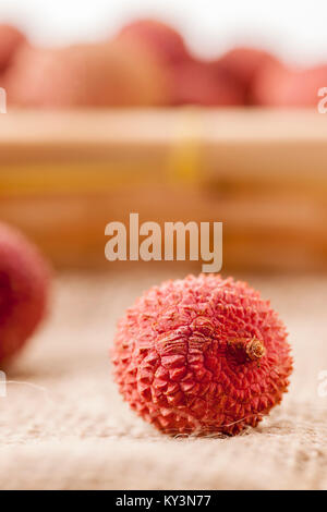 Detail view of a single lychee fruit on rough sackcloth fabric in front of blurred basket with other lychees. Stock Photo
