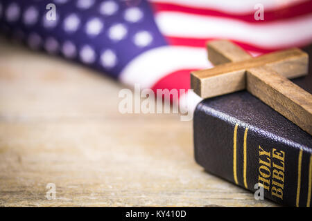 A wooden Christian cross laying on a holy Bible with an American flag background. Stock Photo