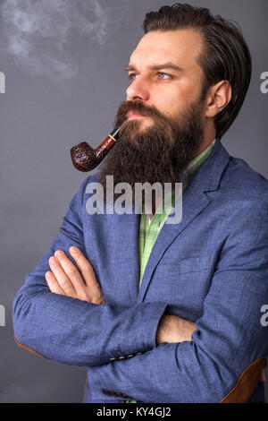 Studio shot of a handsome young man with retro look, arms holded smoking pipe over gray background Stock Photo