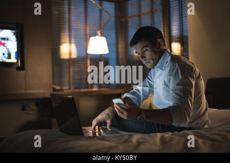 Businessman using laptop and mobile phone in bedroom Stock Photo