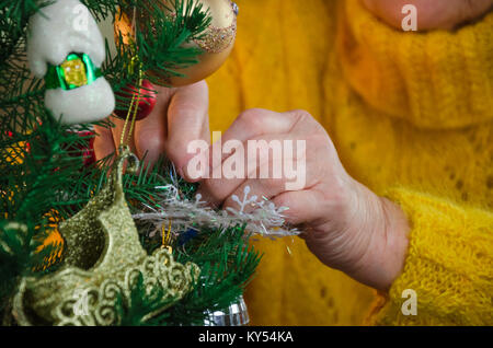 closeup old grandmother's hands decorating fairy tale Christmas tree. senior lady put ball, toy decoration to fir branch. granny in yellow knitted swe Stock Photo