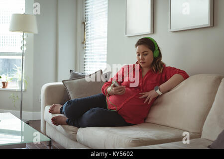 Young pregnant woman sitting on sofa listing to music on her mobile phone Stock Photo