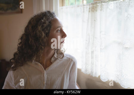 Woman looking through window in living room Stock Photo