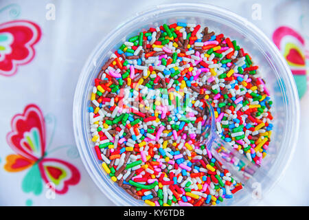 a bowl full of colorful candy sprinkles with a clear plastic spoon in them and a plastic tablecloth Stock Photo