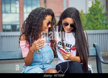Two smiling young women sitting on a city bench in the summer watching something on social media together on a shared pair of headphones Stock Photo