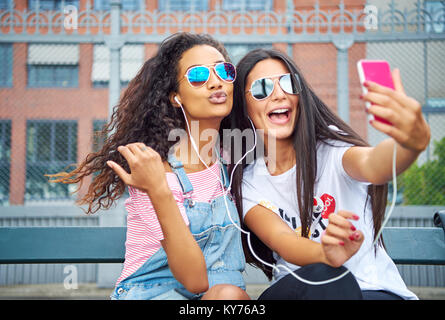 Two young girlfriends smiling and sitting on a city bench listening to music on earphones and taking self portraits together with a smartphone Stock Photo