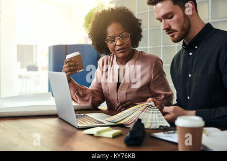 Designers discussing color swatches and using a laptop while sitting together at a table in a modern office Stock Photo