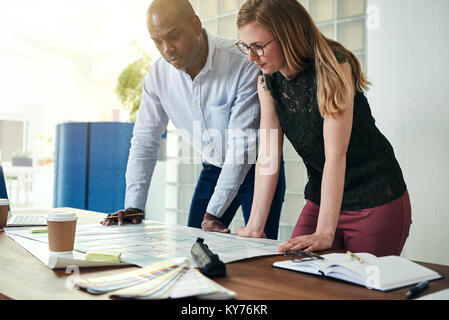 Two focused young architects leaning on a table discussing blueprints together while working in a modern office Stock Photo