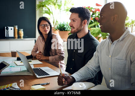 Three diverse work colleagues smiling and talking while sitting together at a boardroom table during a meeting in a modern office Stock Photo