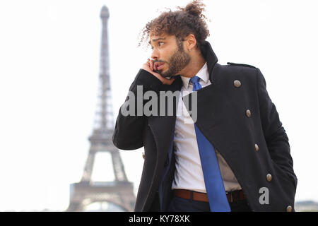 Half Nigerian guy near Eiffel Tower sends messages to girlfriend using modern smartphone in slow motion. Handsome jocund man wears black coat, ring wi Stock Photo