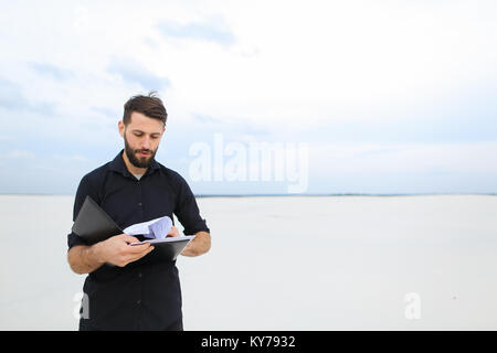 builder man come to deserted place to plan new residential complex, bearded guy standing on sand holding folder with project. Handsome fair-haired con Stock Photo